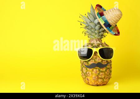 Ananas con cappello sombrero messicano, occhiali da sole e baffi finti su sfondo giallo, spazio per testo Foto Stock