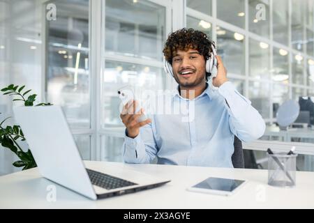 Giovane uomo sorridente che ascolta la musica mentre lavora con il suo smartphone in un ambiente luminoso e moderno. Un concetto di equilibrio tra vita lavorativa e vita privata e tecnologia. Foto Stock