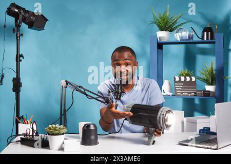 Streamer afro-americano che rivede l'attrezzatura da studio di videografi e registra video. Vlogger che consiglia accessori fotografici agli abbonati dei canali Internet e guarda la fotocamera Foto Stock