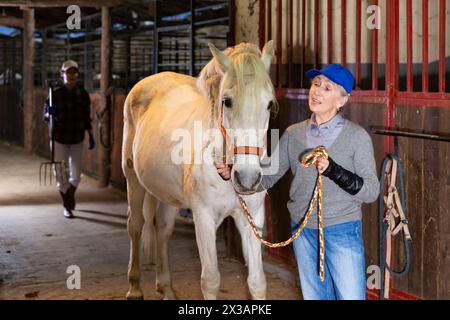 Donna anziana rancher carezzare cavallo bianco dopo l'equitazione Foto Stock