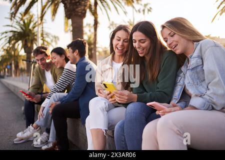 Un gruppo numeroso e diversificato di amici di gen z sorridenti seduti all'aperto utilizzando e guardando telefoni cellulari. Foto Stock
