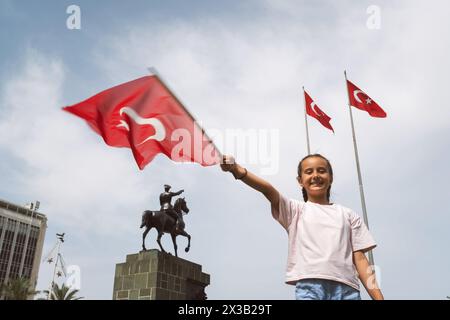 Izmir, Turchia - 23 aprile 2024: Una ragazza gioiosa batte la bandiera turca durante le festività della Festa dei bambini, con la statua di Ataturk a cavallo Foto Stock