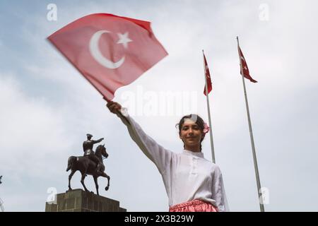 Izmir, Turchia - 23 aprile 2024: Una ragazza gioiosa sventola la bandiera turca durante le festività della Festa dei bambini con un costume tradizionale, con la statua Foto Stock