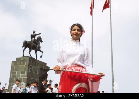 Izmir, Turchia - 23 aprile 2024: Una ragazza gioiosa sventola la bandiera turca durante le festività della Festa dei bambini con un costume tradizionale, con la statua Foto Stock