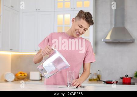 Uomo felice che versa l'acqua dalla caraffa del filtro in vetro al tavolo della cucina Foto Stock