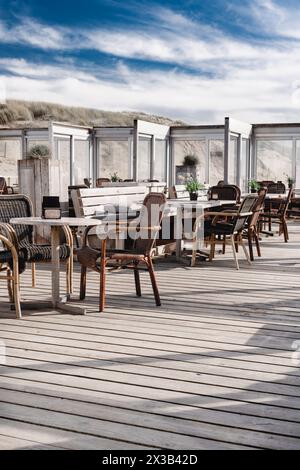 Terrazza del padiglione sulla spiaggia con vista sulle dune di sabbia in una giornata di cielo nuvoloso blu. Terrazza vuota di un padiglione sulla spiaggia, con sedie e tavoli vuoti Foto Stock