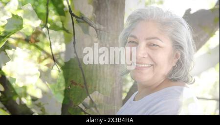 Immagine di macchie di luce e alberi sopra una donna birazziale sorridente in giardino Foto Stock