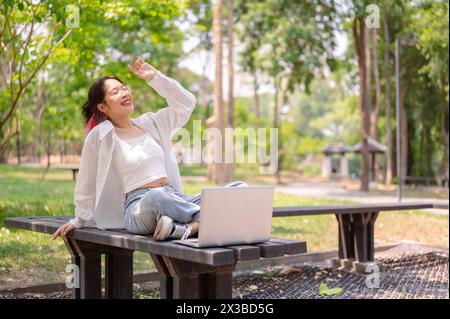 Una giovane donna asiatica spensierata e rilassata, in abbigliamento casual, siede su una panchina in un parco verde in una giornata luminosa, si piega indietro, si allunga le braccia e si gode la natura Foto Stock