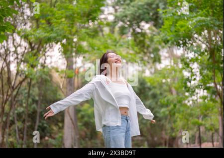 Una giovane donna asiatica spensierata con occhiali e abbigliamento casual si erge con le braccia tese, gli occhi chiusi e un'espressione gioiosa che abbraccia la serenità Foto Stock