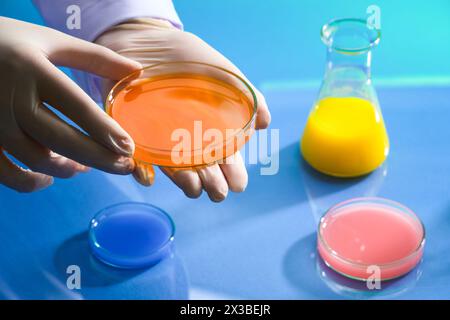 Modello a mano che indossa guanti medici con una piastra Petri di vetro di liquido chimico arancione. Liquido di colore blu, rosa e giallo all'interno di pochi vetri Foto Stock