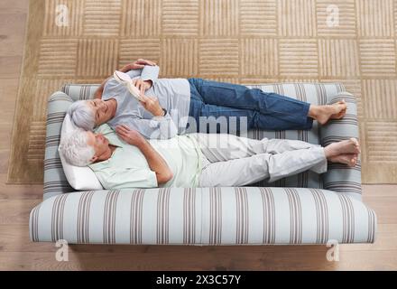 Vista dall'alto, coppia senior e libro da lettura sul divano nel soggiorno per storia, conoscenza e relax a casa. Pensione, anziani e donne sdraiati Foto Stock