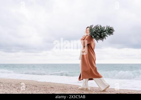 Mare di Natale donna bionda. Ritratto di Natale di una donna felice che cammina lungo la spiaggia e tiene un albero di Natale sulla spalla. Indossa un Foto Stock