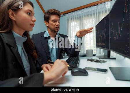 Due commercianti di affari che consultano il mercato azionario dinamico in cambio di investimenti su due schermi di computer, con una collega che ne prende nota Foto Stock