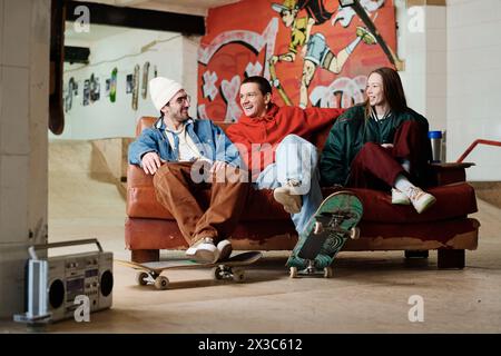 Un gruppo di tre amici maschi e femmine moderni che frequentano lo skatepark seduto sul divano, chiacchierando e ascoltando musica, spazio fotocopie Foto Stock