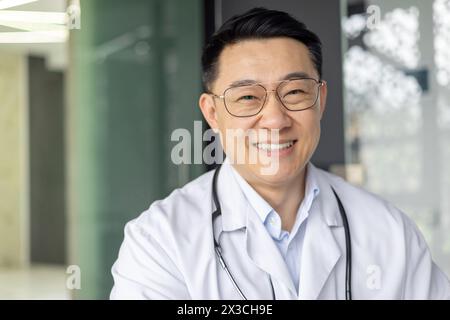 Un ritratto professionale di un medico maschio asiatico maturo, sorridendo con fiducia in un ambiente di clinica medica. Indossa un camice da laboratorio bianco e degli occhiali. Foto Stock