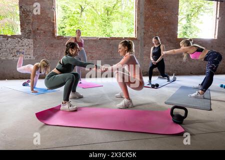 Questa vivace immagine cattura un gruppo di donne impegnate in un allenamento partner all'interno di una palestra industriale. Due donne sono in primo piano, eseguono uno squat mentre si tengono per mano, mostrando il lavoro di squadra e il sostegno reciproco nella loro routine di fitness. In background, altri eseguono esercizi individuali, sottolineando l'importanza della forza personale in un ambiente di gruppo. I mattoni grezzi e gli interni spaziosi riflettono un ambiente di allenamento dedicato, mentre il linguaggio del corpo aperto e il contatto visivo tra i partner evidenziano l'aspetto comune dell'esercizio fisico. Forza collaborativa: Allenamento partner Sessi Foto Stock