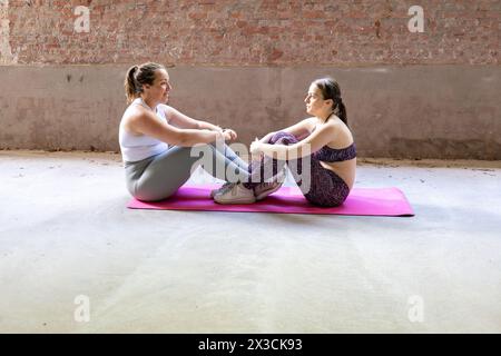 Questa immagine mostra due donne l'una di fronte all'altra, impegnate in un esercizio di seduta assistita su un tappetino condiviso. Si trovano in una spaziosa palestra con pareti in mattoni rustici, indicativa di un ambiente fitness urbano. Sembrano essere a metà movimento, supportandosi reciprocamente le gambe per l'equilibrio e la leva. Il supporto reciproco non è solo fisico ma anche motivazionale, poiché lavorano insieme per raggiungere i loro obiettivi di fitness. Collegamento per l'allenamento dei partner: Esercizio di seduta assistito. Foto di alta qualità Foto Stock