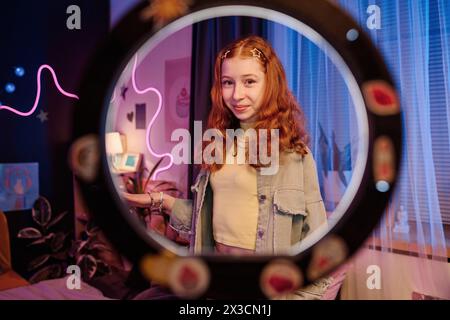 Ritratto di un'elegante ragazza adolescente in piedi nella sua stanza che posa per la fotocamera, attraverso una lampada circolare per scatti medi Foto Stock