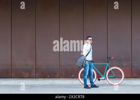 Uomo elegante con occhiali da sole e borsa Messenger accanto a una bici rossa, che effettua una telefonata contro una parete in metallo arrugginito Foto Stock