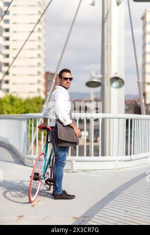 Uomo casual con occhiali da sole, con la sua bici a ingranaggi fissi su un ponte moderno, dietro di lui. Foto Stock