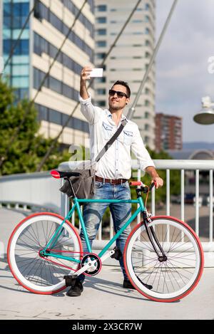 Uomo sicuro di sé con un abito casual scatta un selfie con uno smartphone mentre si appoggia alla sua bici fissa su un ponte urbano. Foto Stock