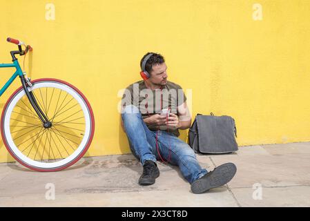 Uomo con un abbigliamento casual con cuffie rosse seduto su una parete gialla brillante accanto a una bicicletta elegante, avvolto nel suo smartphone con una borsa nelle vicinanze Foto Stock