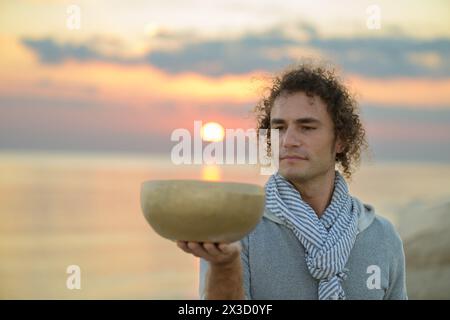 Uomo che tiene in mano una ciotola tibetana che canta contro il mare al tramonto Foto Stock