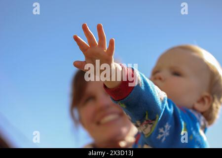 Bambino che si avvicina sotto un cielo blu, madre sorride dietro Foto Stock