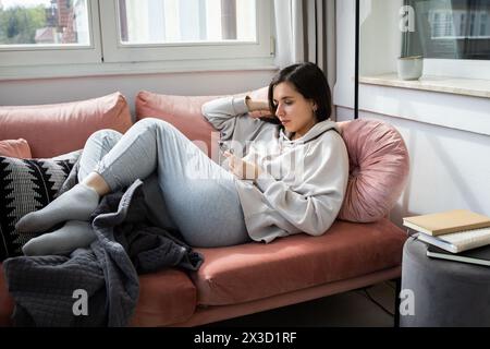 Giovane donna sdraiata su un pullman con abiti comodi e che scorre al telefono Foto Stock