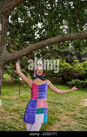 Una bella giovane donna con un abito vivace e occhiali da sole si staglia sotto un albero, crogiolandosi nella brezza estiva. Foto Stock