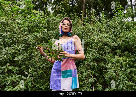 Una giovane donna vivace con un abito colorato e occhiali da sole si erge di fronte a un cespuglio, tenendo delicatamente in mano una pianta. Foto Stock