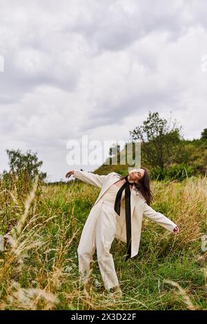 Una giovane donna sta in piedi in un campo, braccia allungate, indossa abiti bianchi, sentendo la brezza estiva e abbracciando la bellezza della natura. Foto Stock