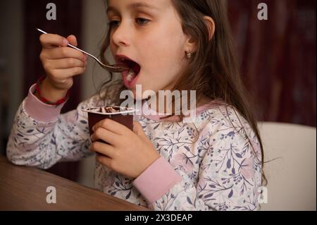 Ragazzina europea che stuzzica con un gustoso yogurt a casa. Ritratto autentico di un bambino di 6 anni, gustando il gusto del cioccolato vega Foto Stock