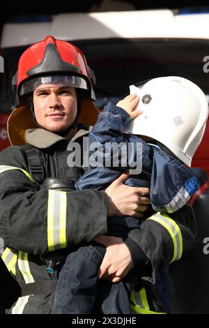 Ritratto di un pompiere che indossa proiettili e casco rosso, tiene il bambino tra le braccia Foto Stock