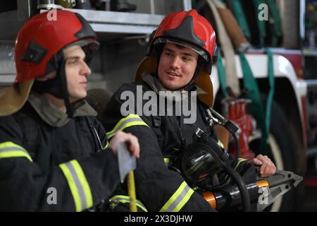 Due vigili del fuoco in tute protettive e un casco rosso per proteggere la testa con l'attrezzo nelle mani seduti contro il motore antincendio Foto Stock