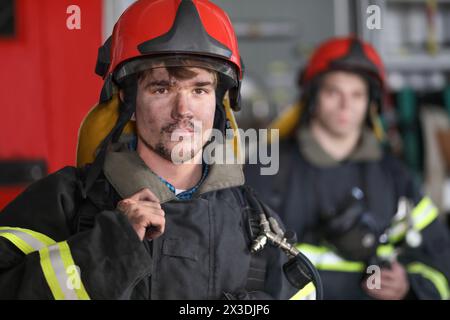 Ritratto di due eroici pompieri in tuta protettiva e casco rosso, secondo pompiere fuori fuoco, primo piano Foto Stock