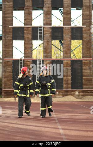 Due vigili del fuoco in equipaggiamento e caschi rossi nell'area di prova contro la torre antincendio Foto Stock