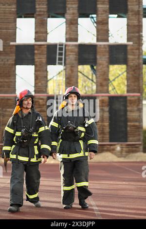 Due vigili del fuoco in equipaggiamento e caschi rossi vanno nell'area di prova di addestramento contro la torre antincendio Foto Stock