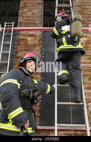 due vigili del fuoco in equipaggiamento e caschi rossi sul sito di prova, uno di loro sale le scale Foto Stock