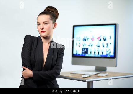 Una giovane donna con una giacca nera sta con le mani agganciate sullo sfondo della scrivania del computer e del computer Foto Stock