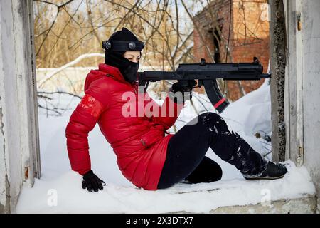 Donna in giacca rossa e maschera da sci che mira con pistola da gioco seduta a terra all'aperto il giorno invernale. Foto Stock