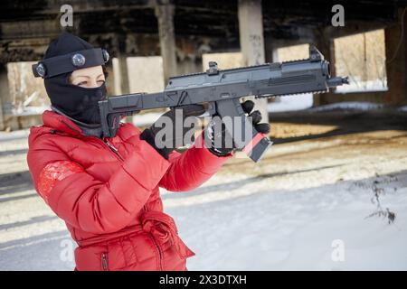 Donna in giacca rossa e maschera da sci che mira con pistola da gioco all'aperto il giorno invernale. Foto Stock