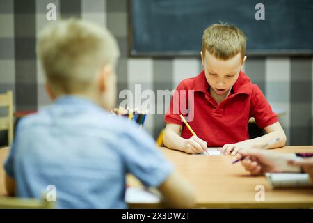 I bambini disegnano con le matite sedute alla scrivania in classe e si concentrano sul ragazzo con la camicia rossa. Foto Stock