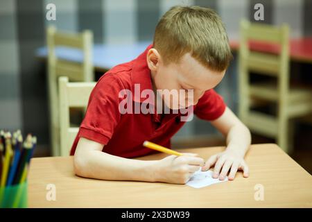 Il bambino con la camicia rossa disegna con una matita su un foglio di carta seduto alla scrivania in camera. Foto Stock