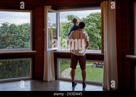 Padre sta alla finestra aperta sulla veranda, tenendo il figlio sulle braccia, vista posteriore. Foto Stock
