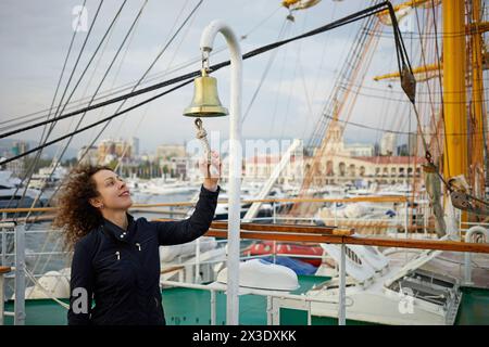 Donna con capelli ricci in una giacca scura che aggancia la campana sul ponte della nave. Foto Stock