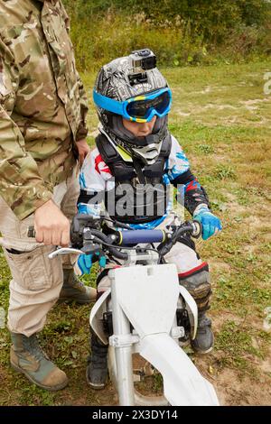 Il bambino, equipaggiato e vestito per le gare motociclistiche, è seduto sulla moto e l'uomo tiene la barra della maniglia. Foto Stock