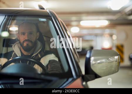 L'uomo barbuto siede al posto di guida in auto nel parcheggio sotterraneo. Foto Stock