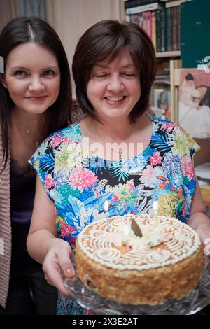 Due donne stanno nel soggiorno tenendo una torta di compleanno con candele accese. Foto Stock