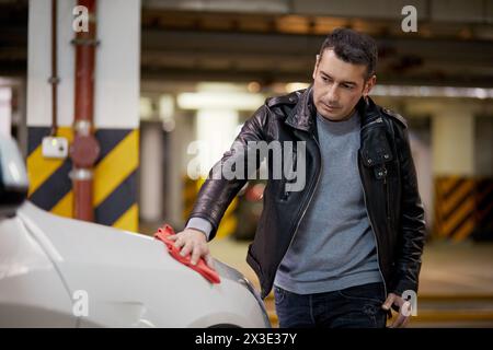 L'uomo lucida la superficie del cofano di auto bianca con un pezzo di tessuto nel parcheggio sotterraneo. Foto Stock